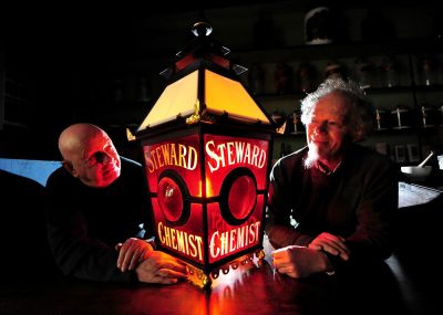 Two men lean either side of a large Victorian era Chemist-shop lamp which featured red glass and has the words "Steward Chemist".
