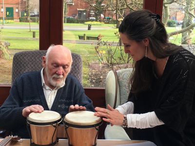 An elderly gentleman plays small drums while a facilitator guides him.