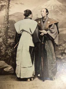 Photograph, c.1870s. Two samurai with swords pose in front of a painted backdrop. By the time this photograph was taken the samurai way of life was rapidly changing, and giving way to new fashions and short haircuts. The photograph was likely produced for a western audience, with the focus very much on the elaborate traditional clothing. Reproduced with kind permission of the Museum of Royal Worcester.